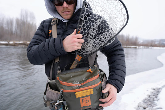 Fishing net with white mesh head and brown handle, held outdoors beside waist pack