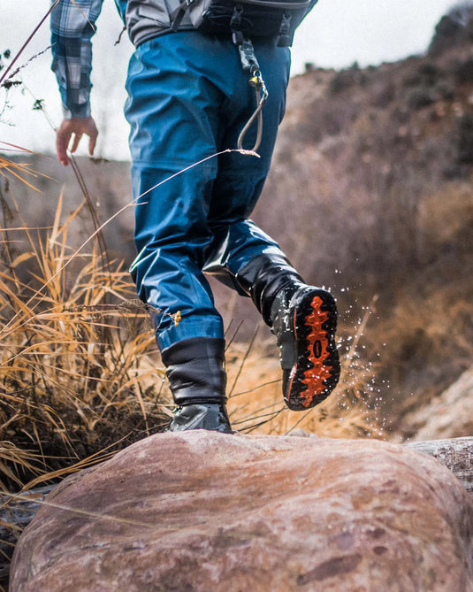 Lace-up rubber-sole boot stepping near rocks, dark upper with orange outsole.
