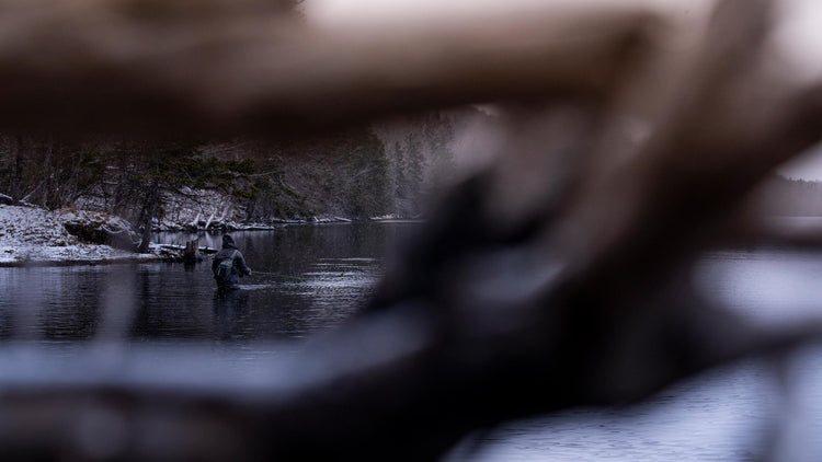 Person fishing from a wooden dock into a lake with trees and snow in the background